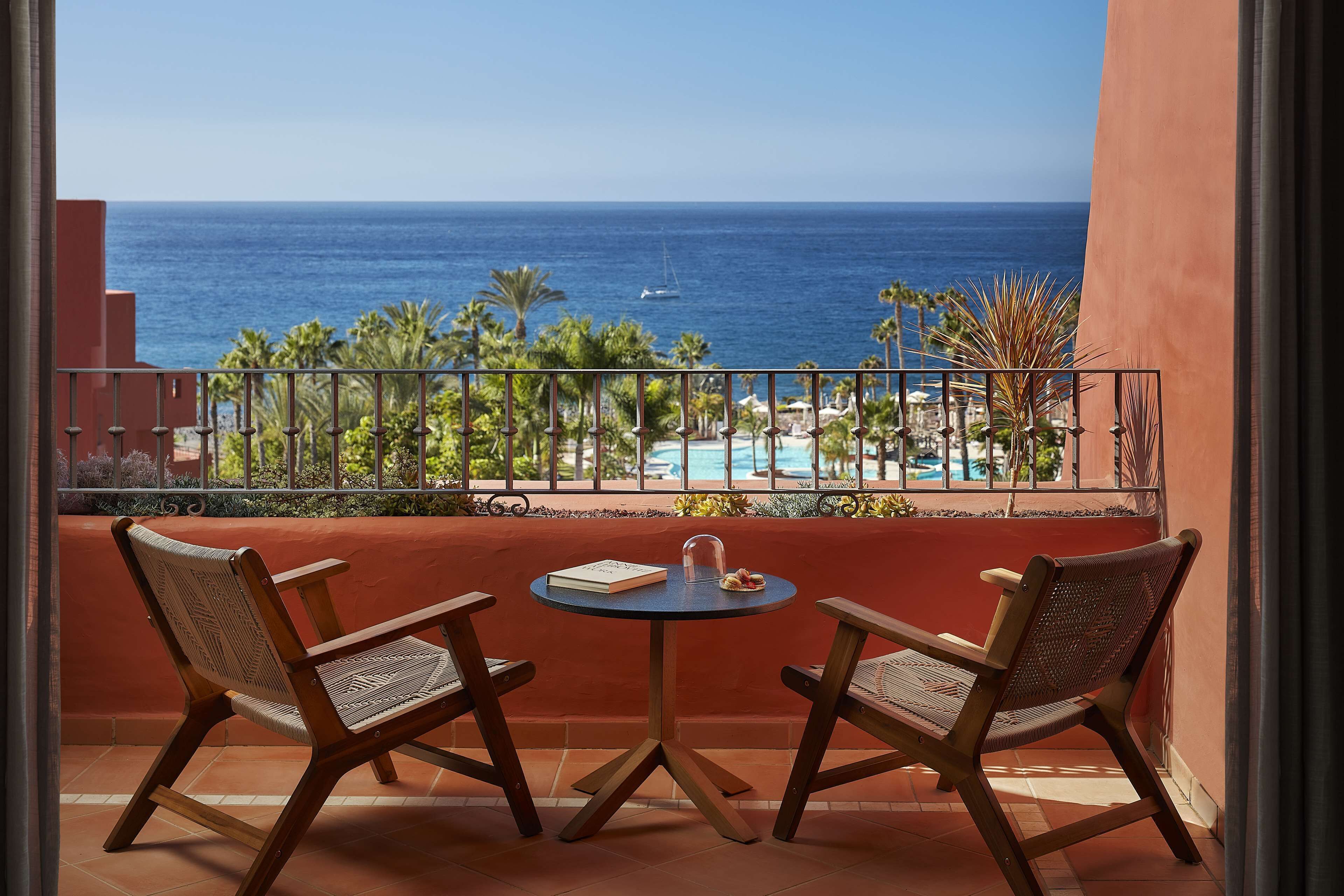A balcony with two chairs and a table, overlooking a beach and ocean.