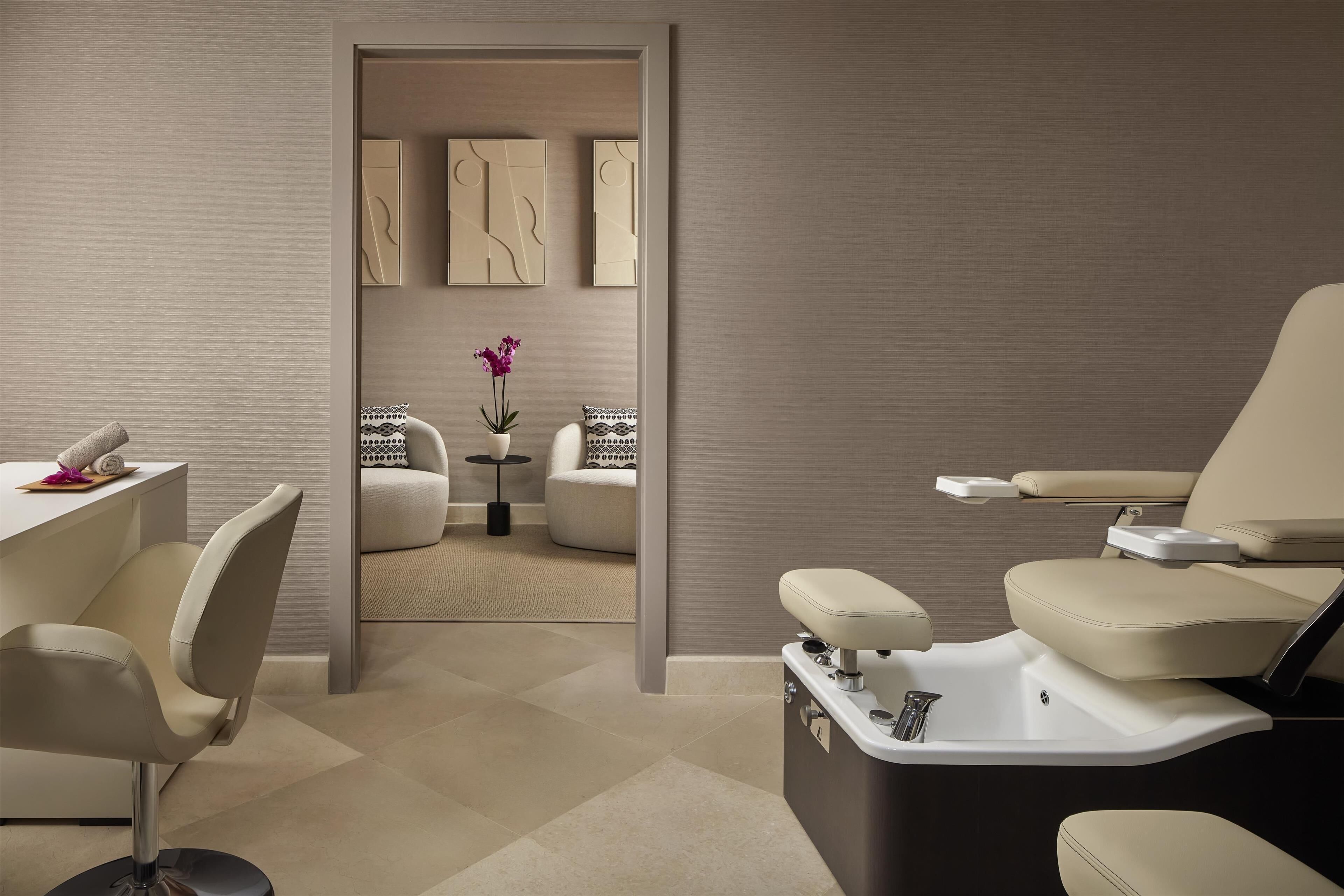 A modern spa room with a massage table, a chair, and a small table with a vase of flowers.