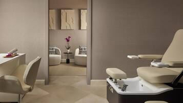 A modern spa room with a massage table, a chair, and a small table with a vase of flowers.