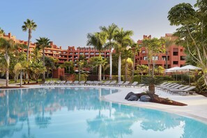 A pool area with lounge chairs and palm trees, surrounded by red buildings. - Tivoli la Caleta Resort (Adeje)