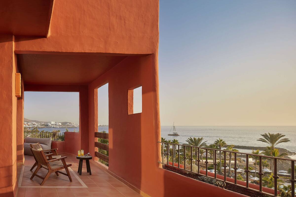 A balcony with a view of the sea, a wooden chair, a small table with a drink, and a railing with umbrellas.