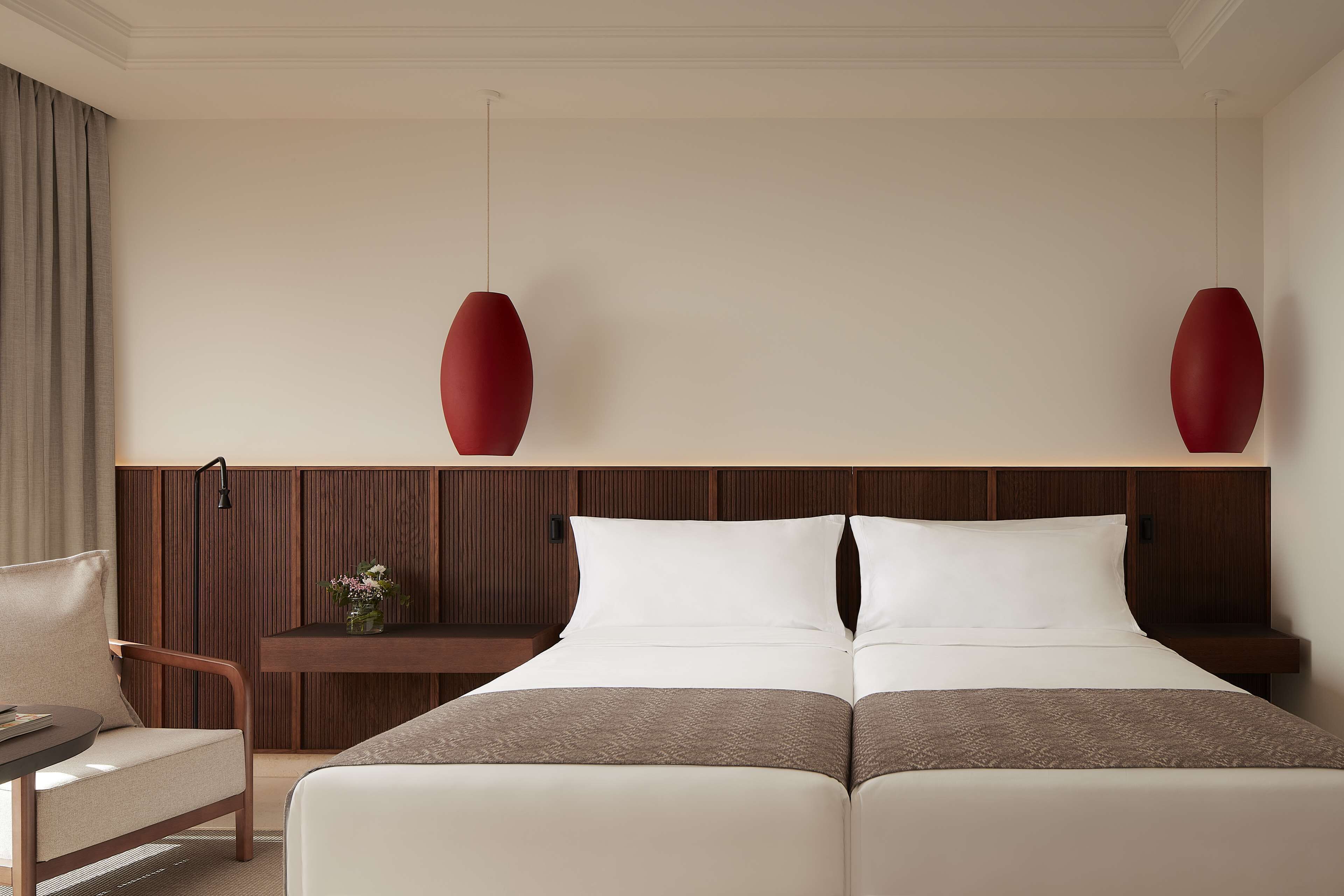 Une chambre d’hôtel avec un lit double, une tête de lit en bois, une petite table sur laquelle est posé un vase de fleurs, et deux lampes suspendues rouges.