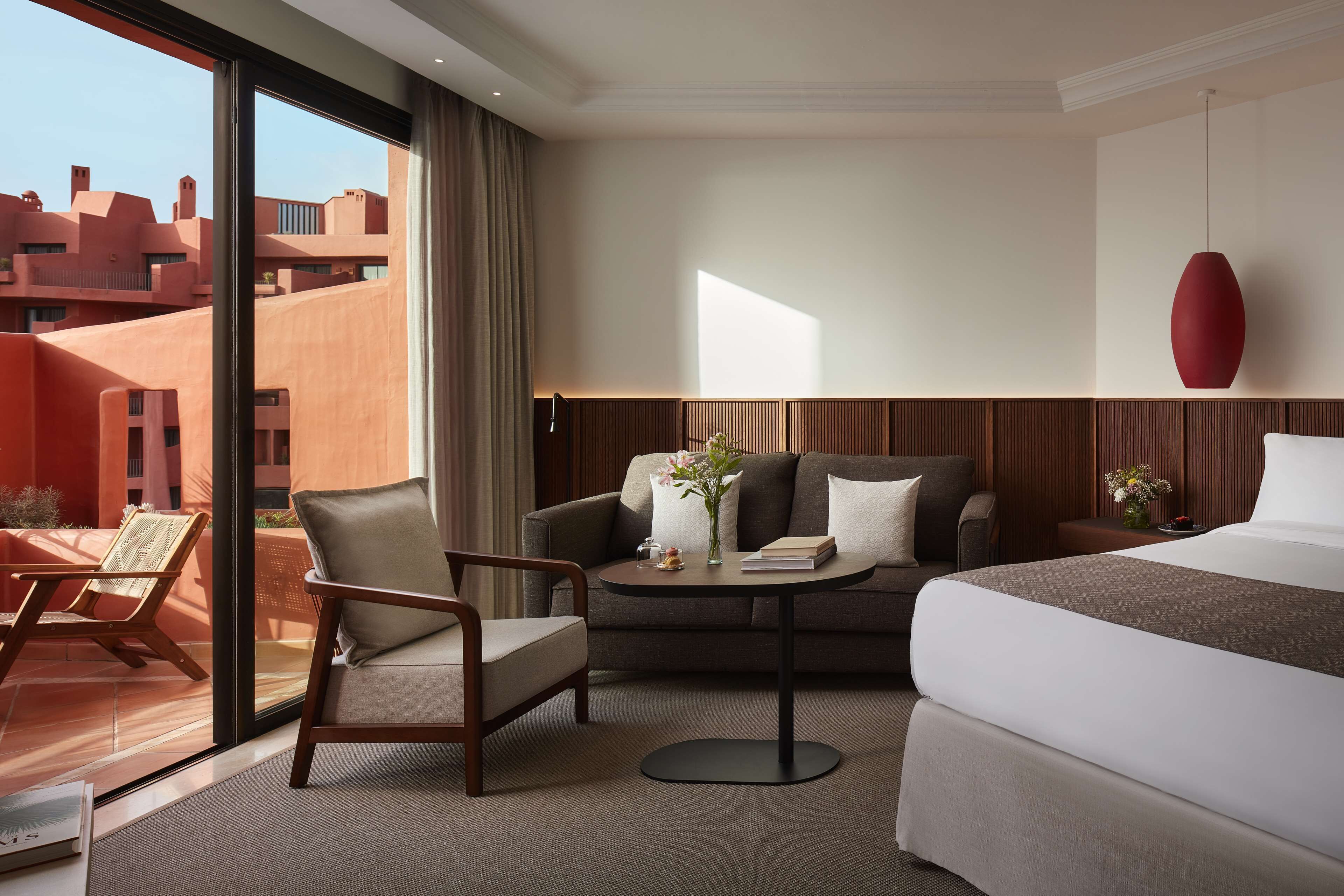 Quarto de hotel com cama, sofá, poltrona, pequena mesa com um vaso e um livro, e vista para edifícios.