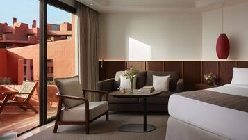 Quarto de hotel com cama, sofá, poltrona, pequena mesa com um vaso e um livro, e vista para edifícios.