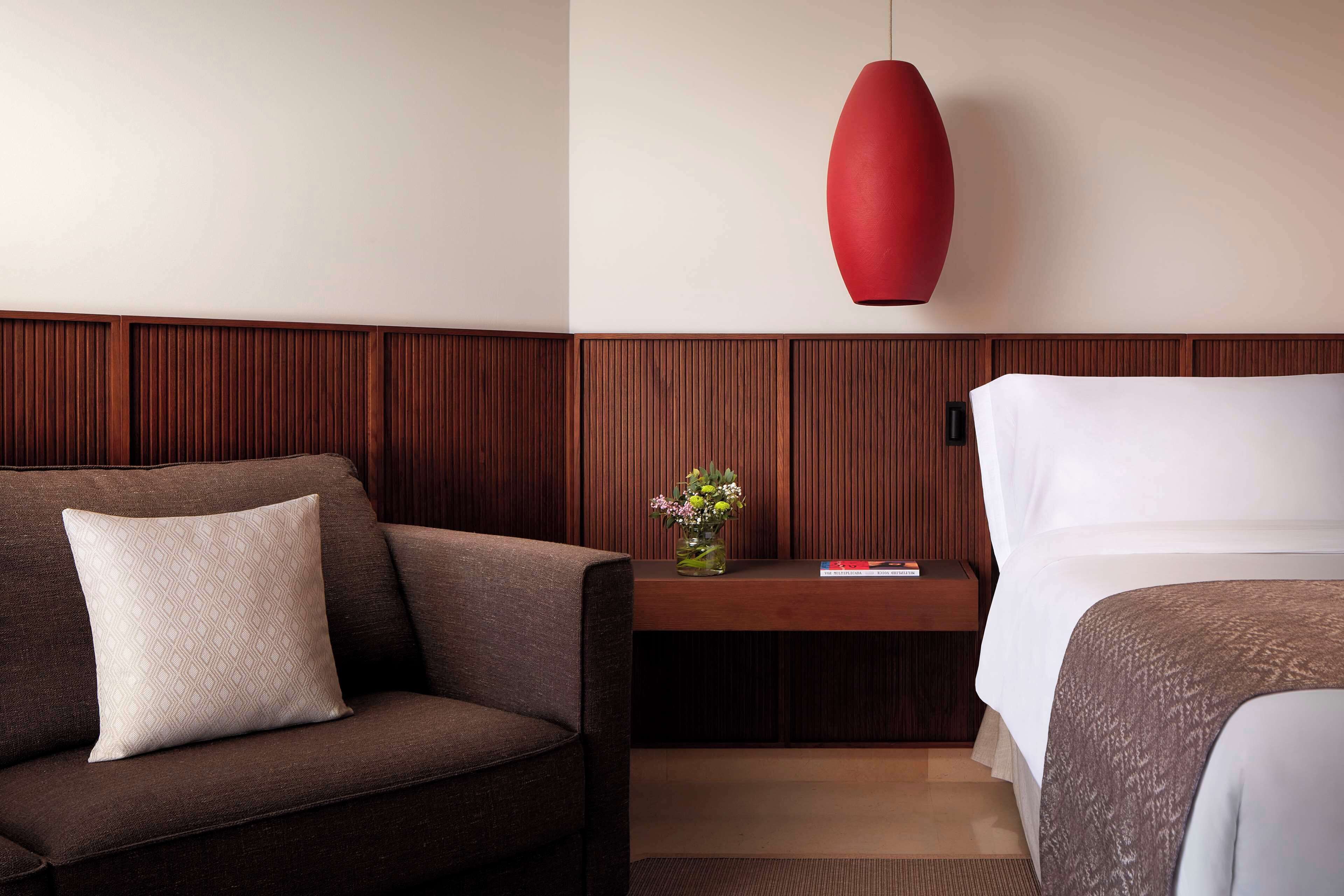 Habitación de hotel con sofá marrón, mesita de noche de madera con un florero y una lámpara colgante roja, además de una cama con ropa blanca.