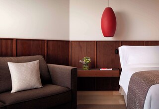 A hotel room with a brown sofa, a wooden nightstand with a vase of flowers, a red pendant light, and a bed with white bedding.