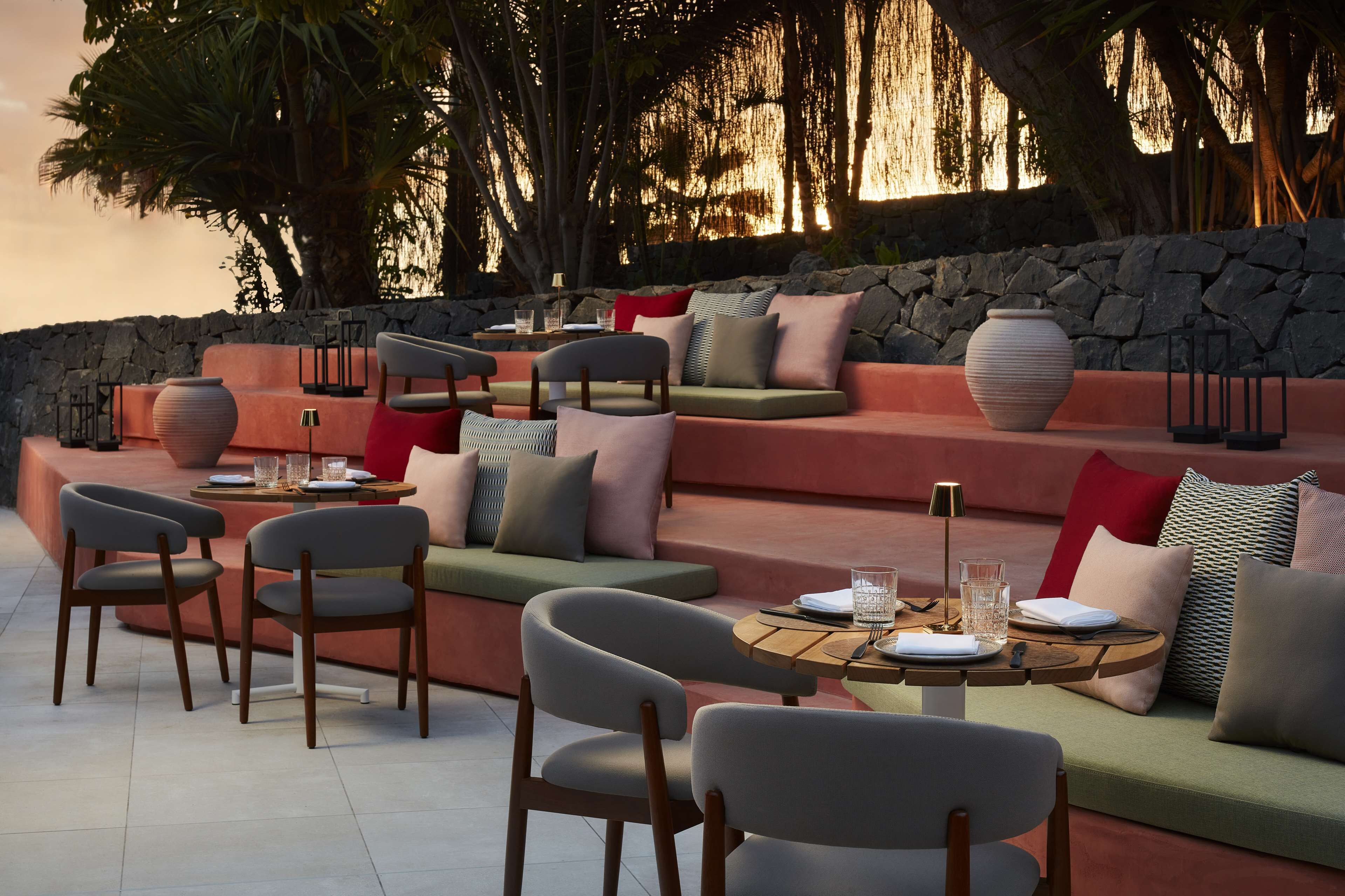 Un área de estar exterior moderna con bancos acolchados, sillas y una mesa redonda dispuesta para comer.