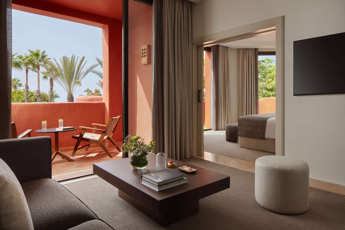 A modern hotel room with a balcony, a sofa, a coffee table, and a view of palm trees.