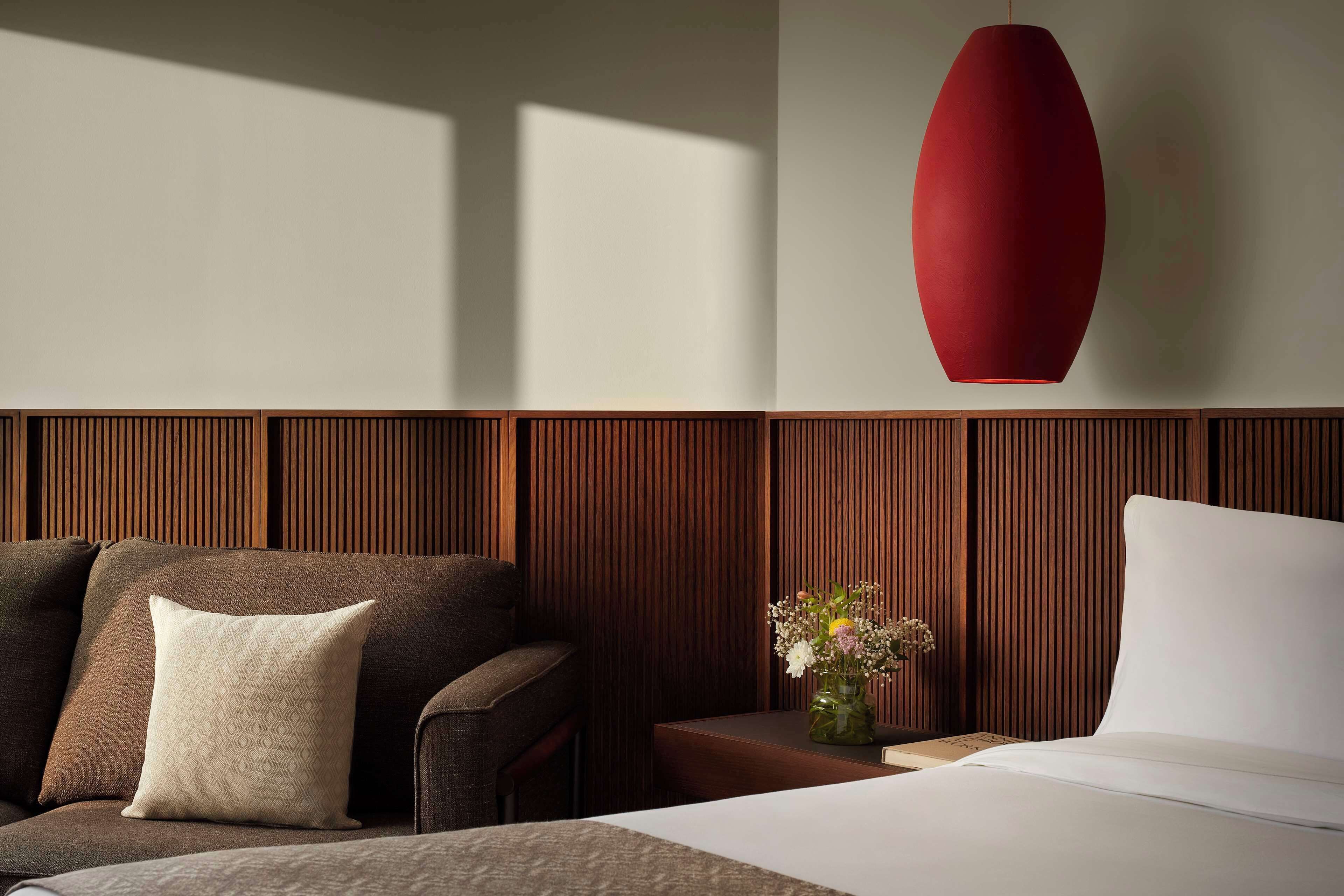 Habitación de hotel moderna con pared de paneles de madera, una lámpara colgante roja, un sofá con un cojín estampado, una mesita con un florero y una cama con ropa de cama blanca.