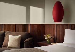 A modern hotel room with a wooden panel wall, a red pendant light, a sofa with a patterned pillow, a small table with a vase of flowers, and a bed with white bedding.