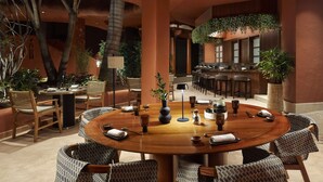 A spacious indoor dining area with a round wooden table surrounded by patterned chairs, a bar area with stools, and potted plants.
