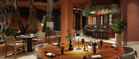 A spacious indoor dining area with a round wooden table surrounded by patterned chairs, a bar area with stools, and potted plants.