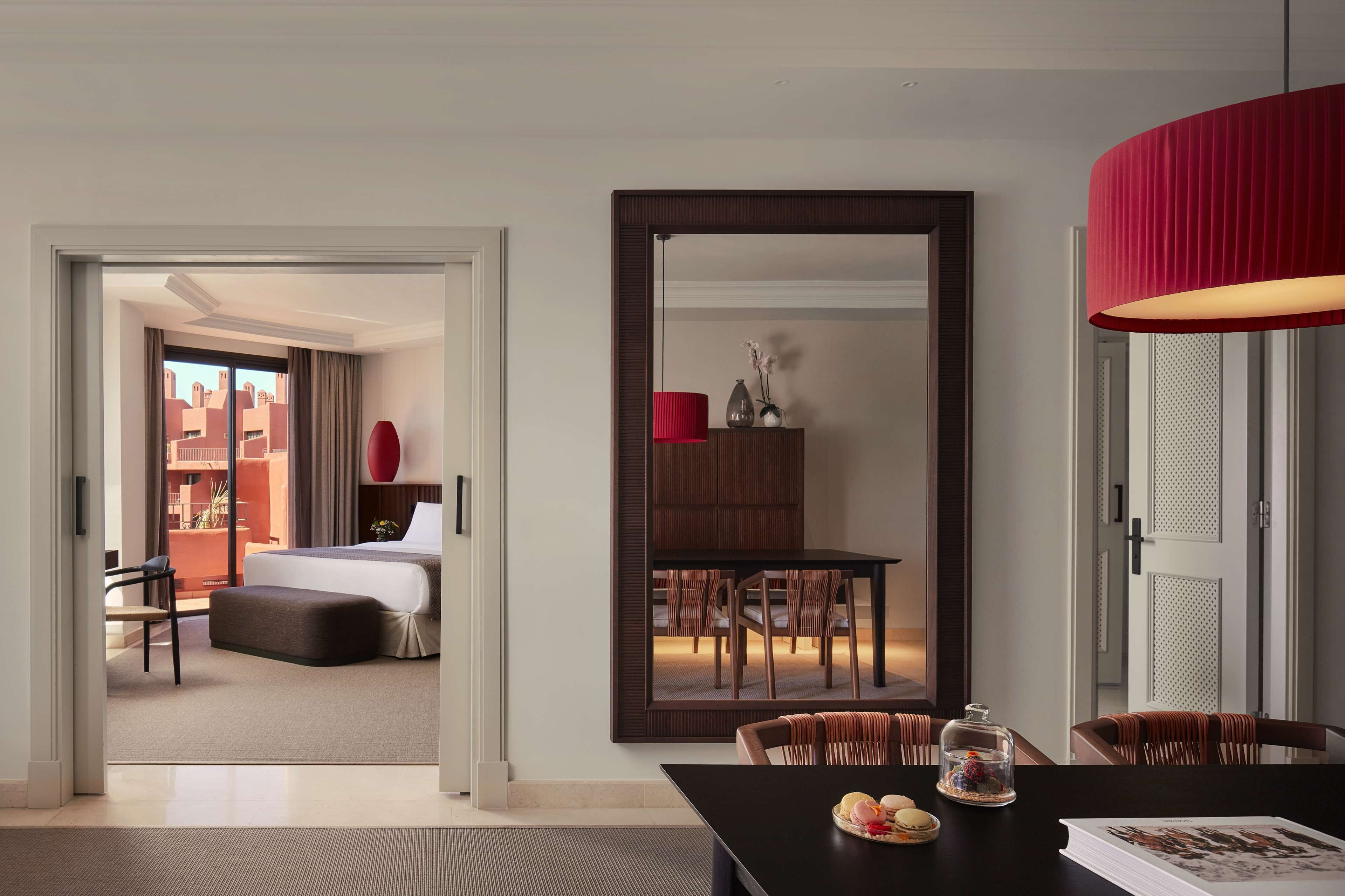 Habitación de hotel moderna con una cama grande, un área de comedor con una lámpara roja y vistas a la ciudad por la ventana.