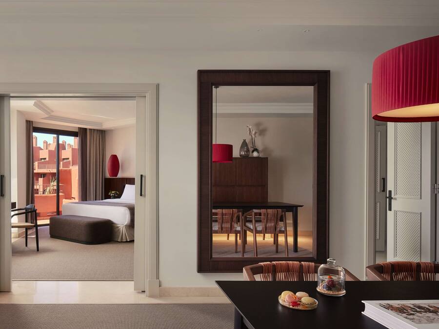 A modern hotel room with a large bed, a dining area with a red lamp, and a view of the city through the window.