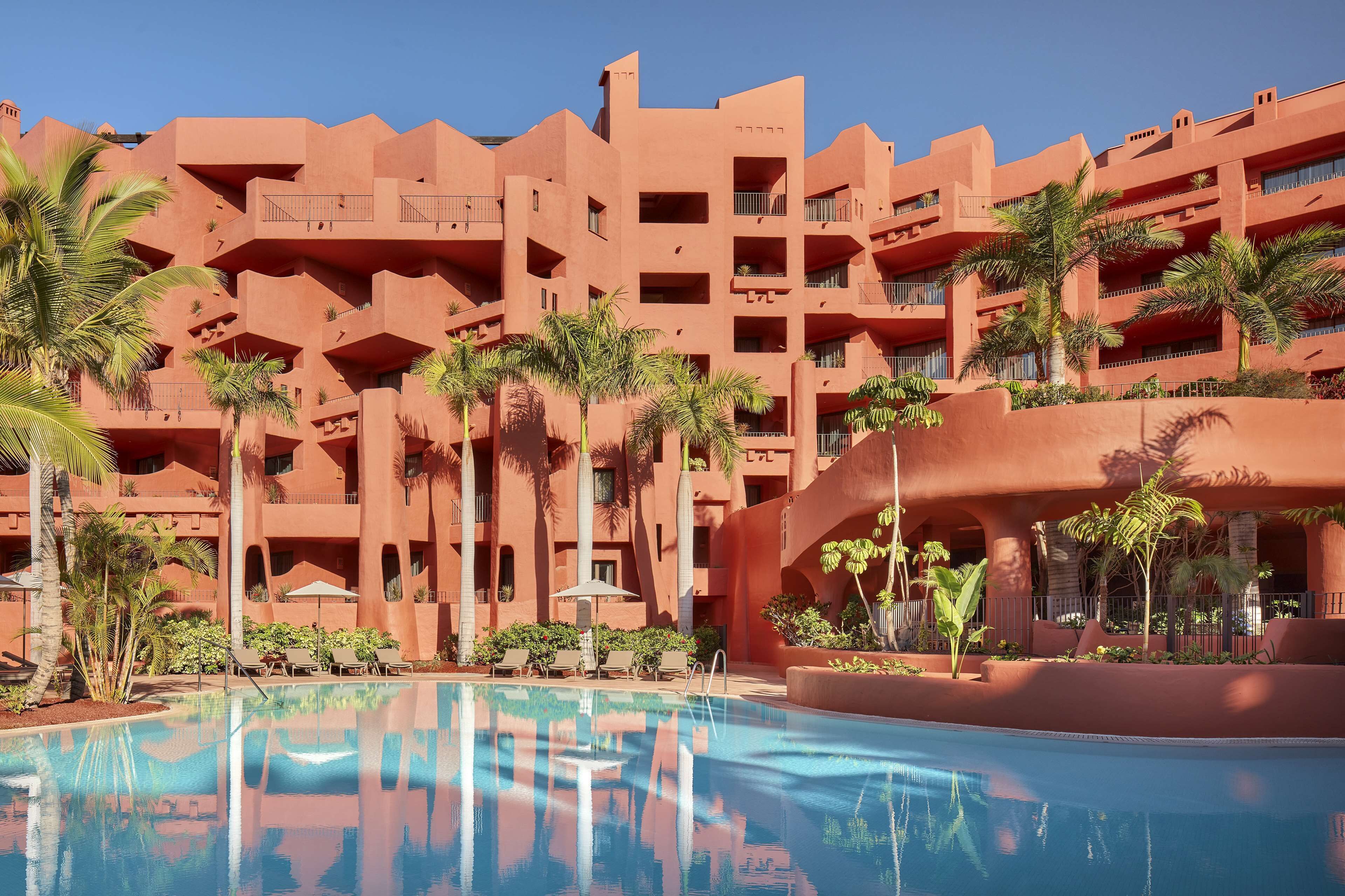 A hotel with a pool, palm trees, and a red building facade.