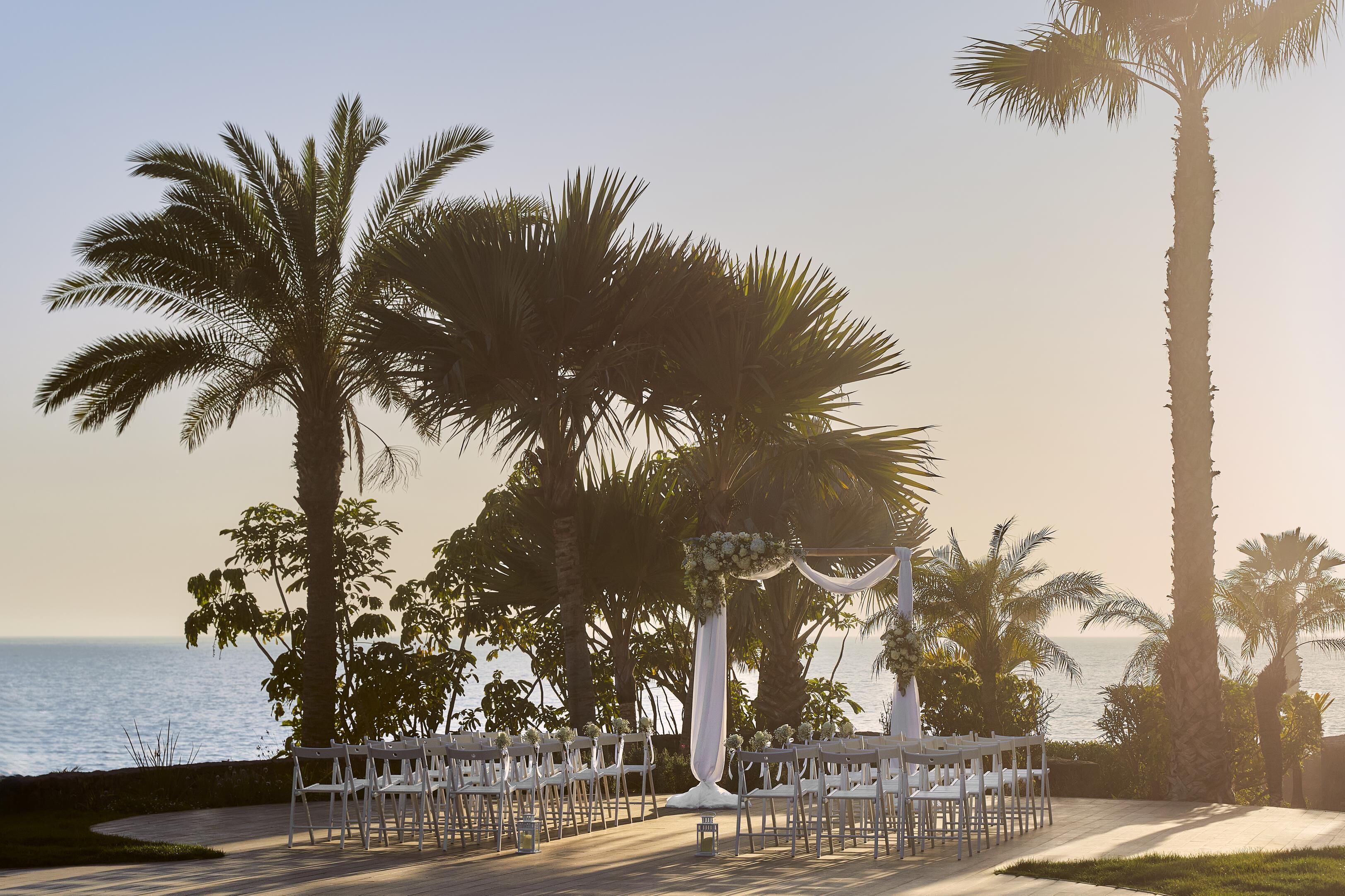 Un lugar para bodas al aire libre con sillas blancas dispuestas en filas, palmeras y vista al mar.