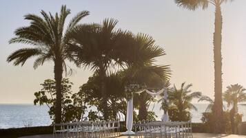 Un lugar para bodas al aire libre con sillas blancas dispuestas en filas, palmeras y vista al mar.