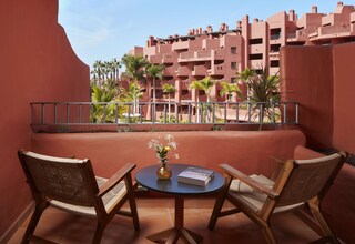 A balcony with two wicker chairs, a small round table, and a book. The balcony overlooks a building with red walls and balconies, and palm trees are visible in the background.