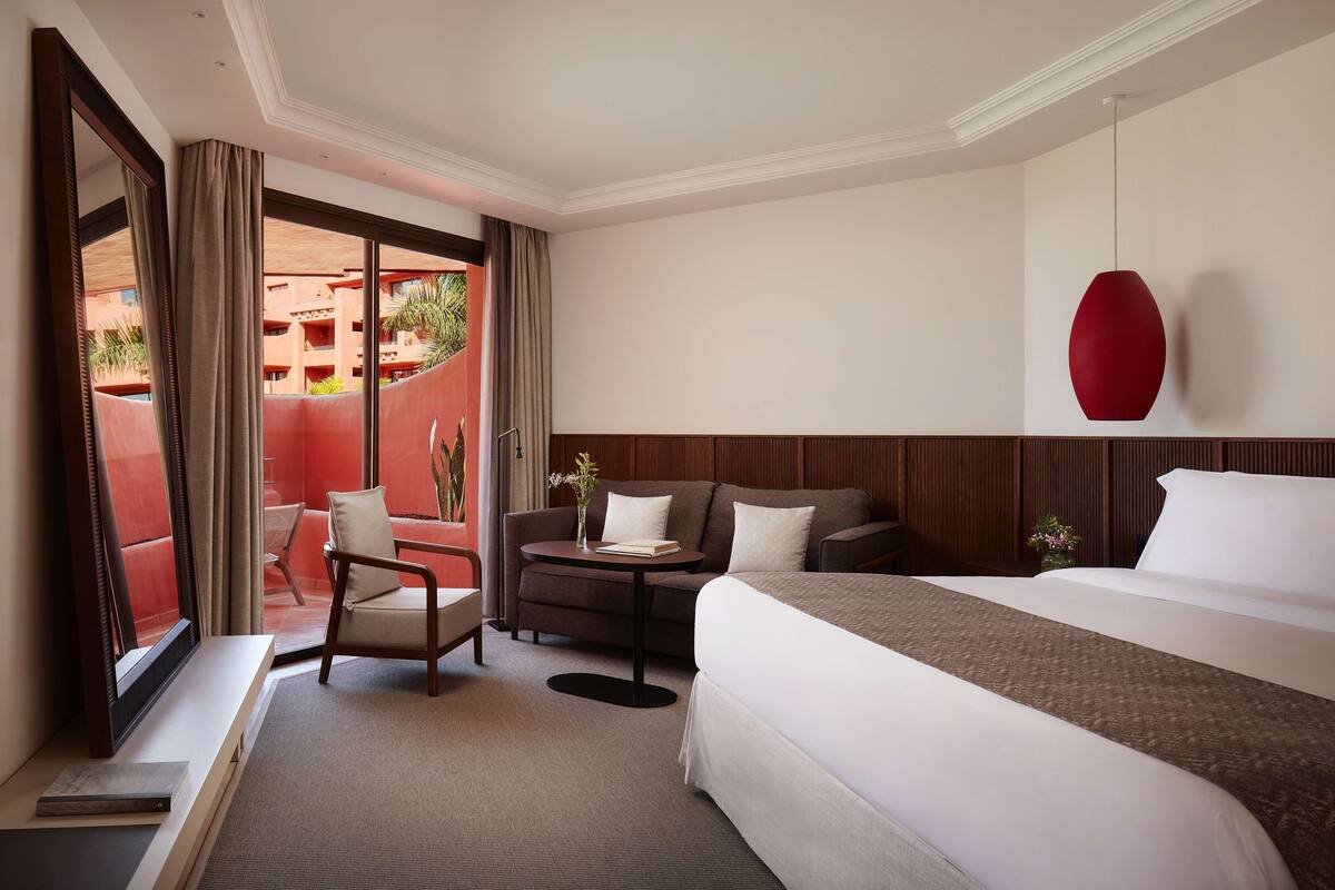 A hotel room with a bed, a desk, a chair, a small table, and a large window.