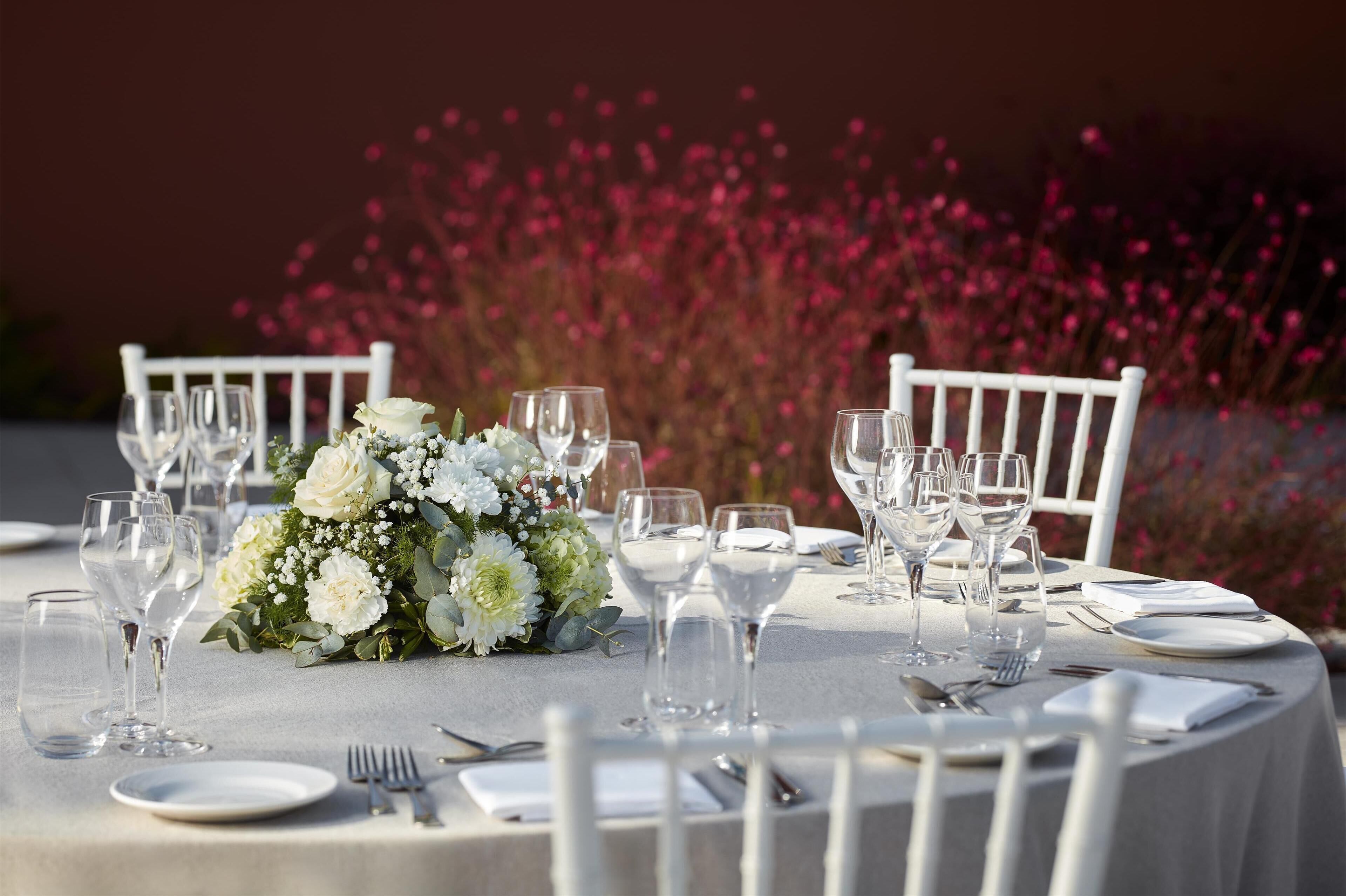 Una mesa redonda dispuesta con sillas blancas, cristalería transparente y un centro de mesa floral.