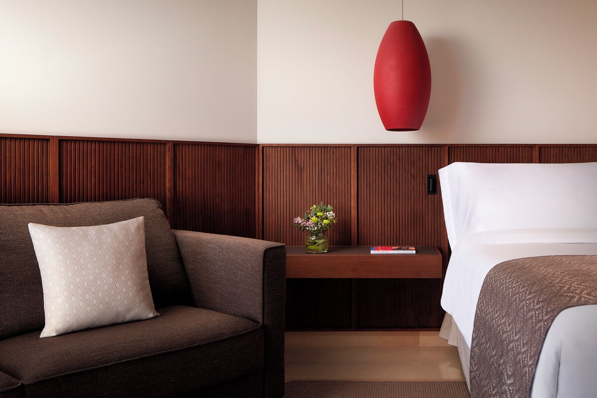A hotel room with a wooden panel wall, a red pendant light, a small bedside table with a vase of flowers, a sofa with a cushion, and a bed with a patterned blanket.