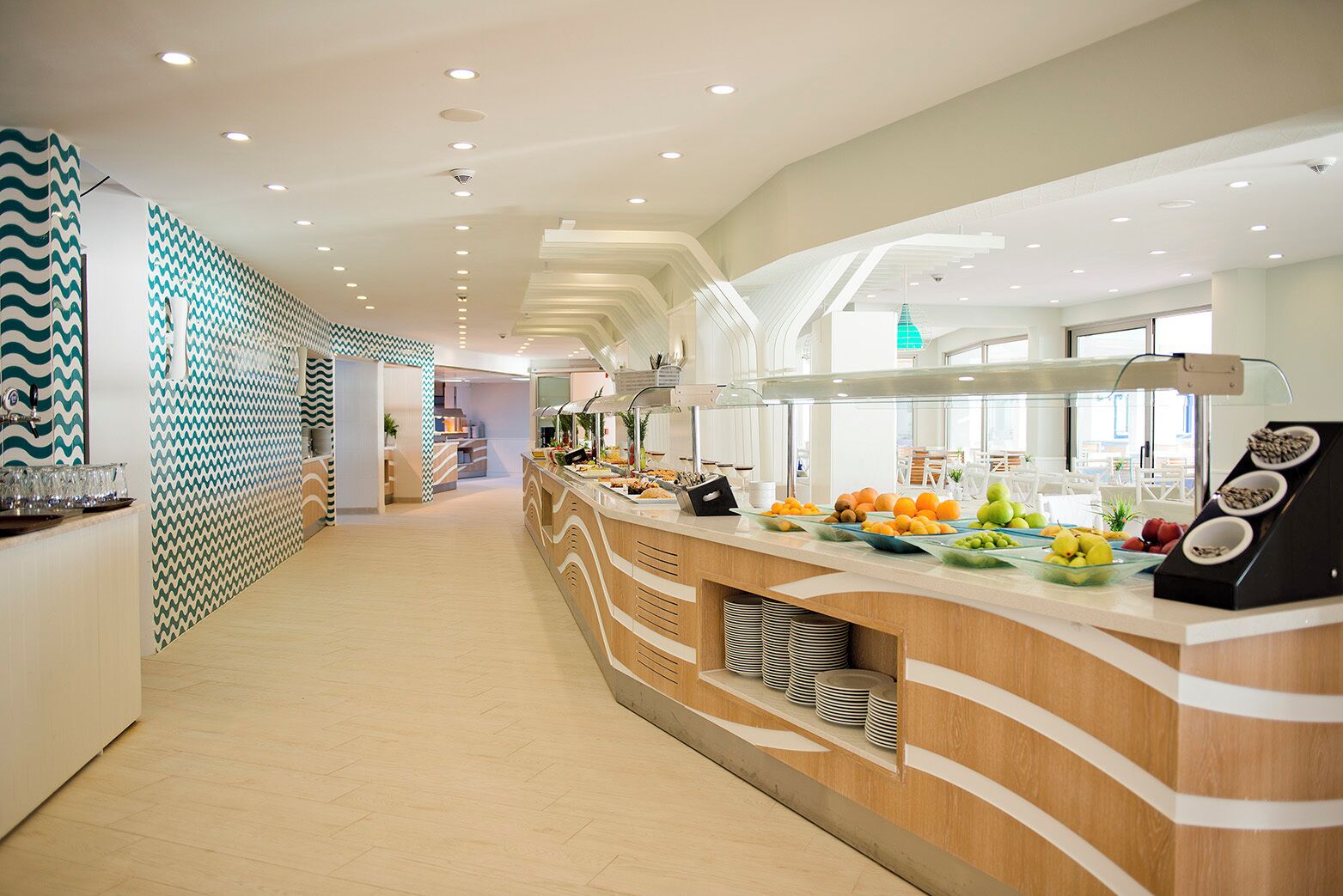 Um hall de hotel moderno com um balcão de recepção extenso, parede estampada e área de buffet com frutas e lanches.