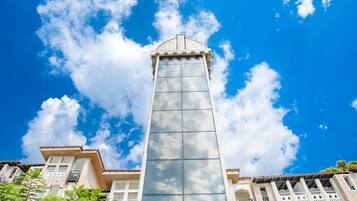 Un edificio alto y moderno con fachada de vidrio y techo plano, rodeado de vegetación y otros edificios.