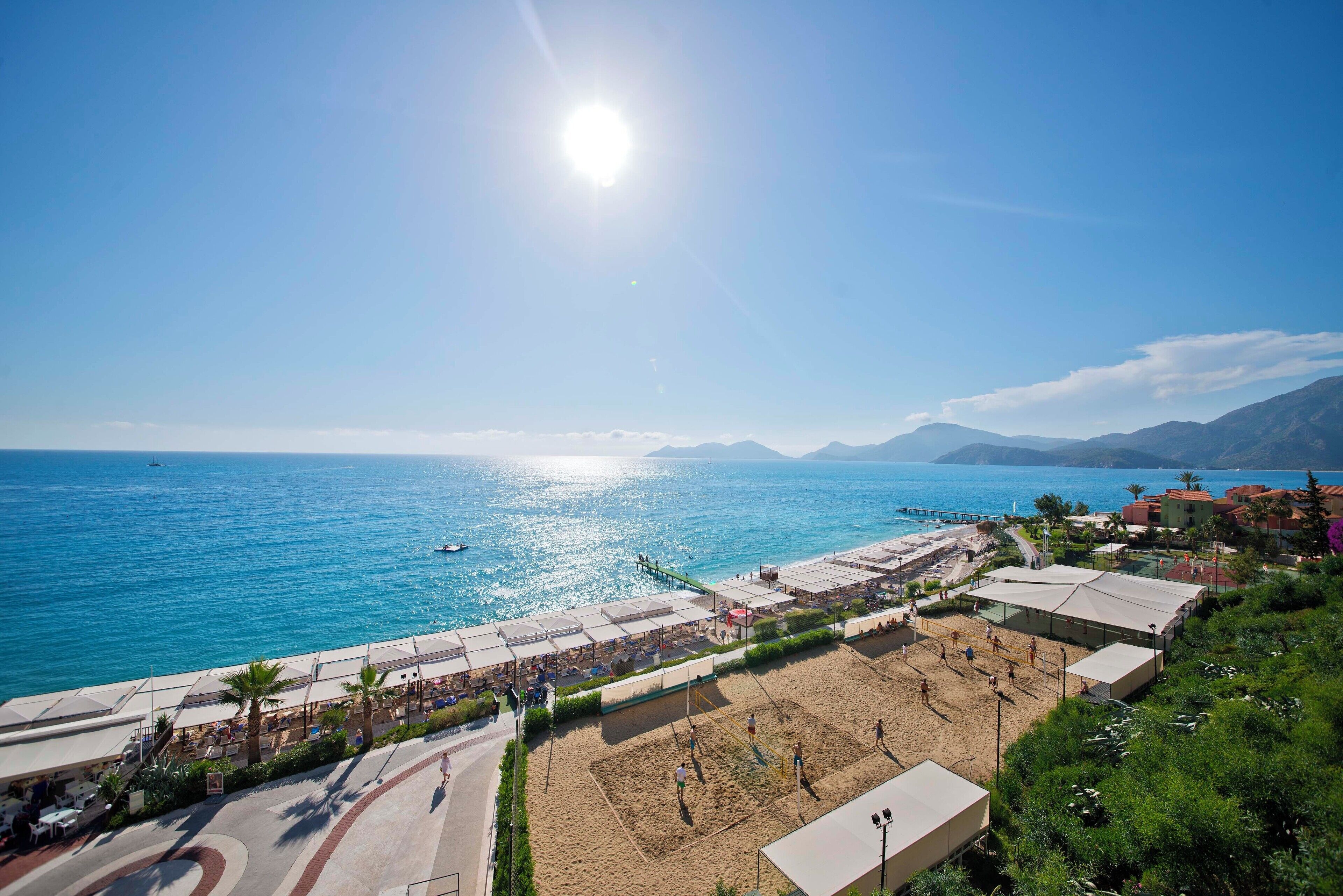 A beachside area with a volleyball court, umbrellas, and a view of the sea and mountains.