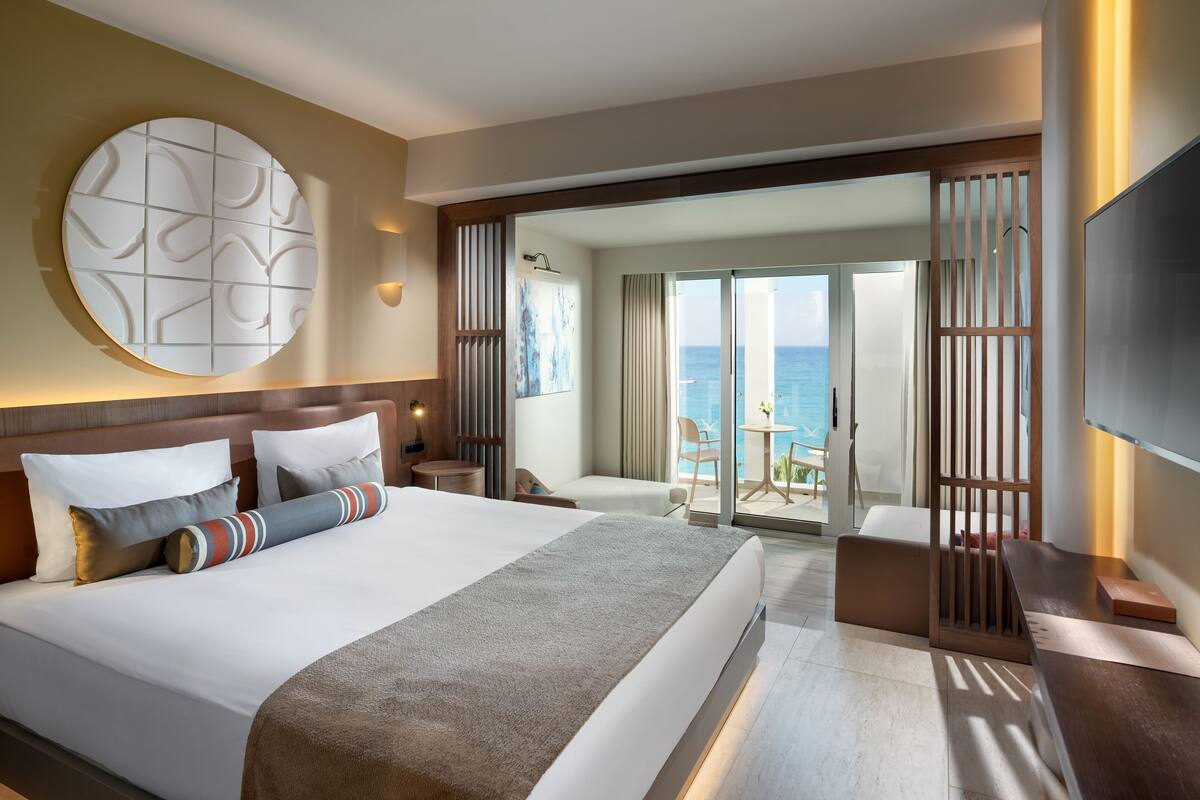 A modern hotel room with a large bed, a TV, a balcony with a view of the sea, and a decorative wall piece.