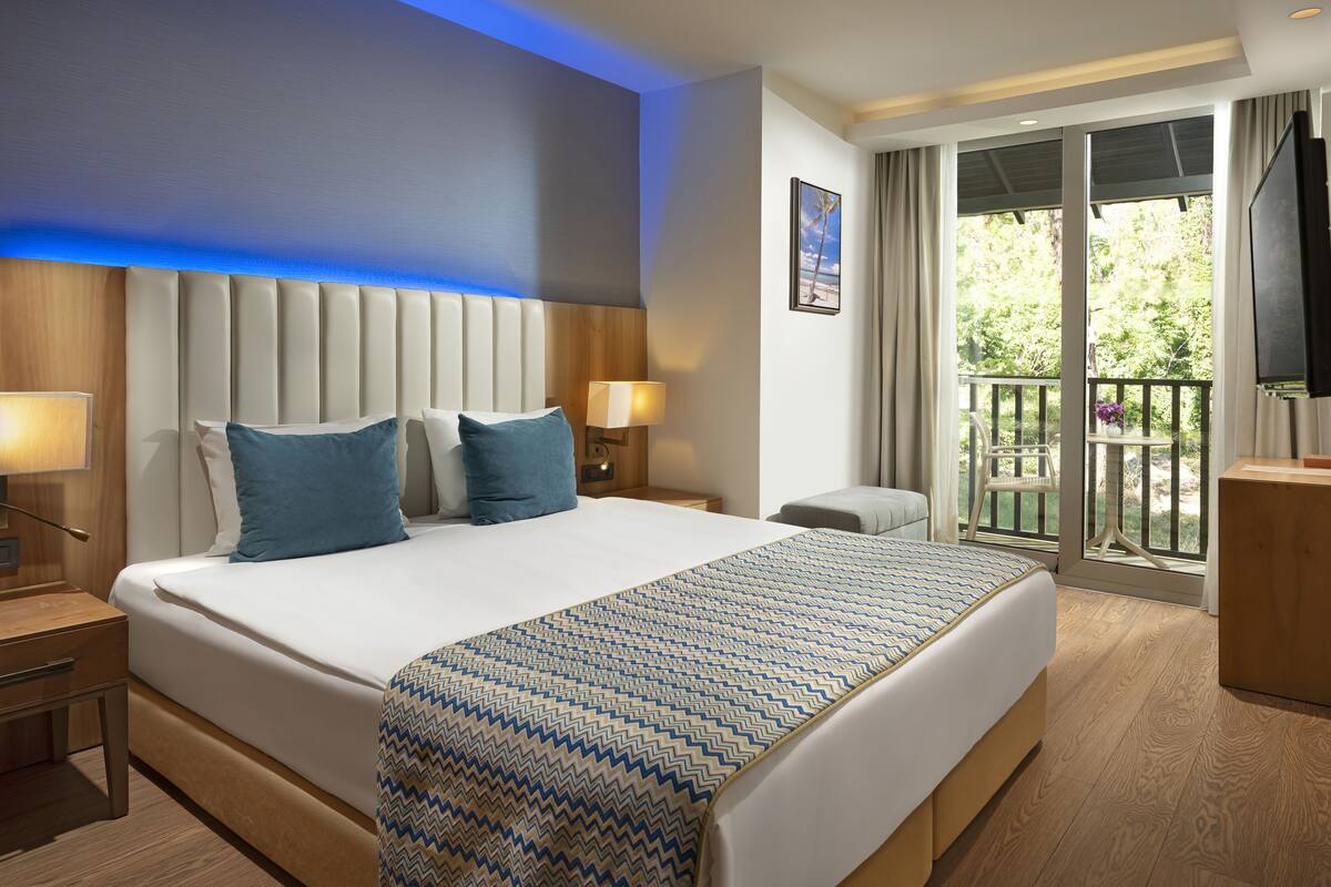 A hotel room with a large bed, two bedside tables, a nightstand, a TV, and a balcony with a view of greenery.
