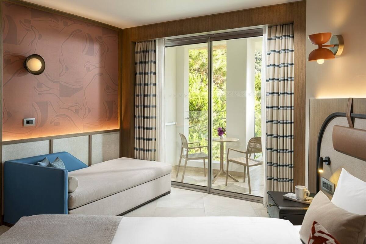 A hotel room with a bed, a sofa, a small table with a chair, a nightstand, and a sliding glass door leading to an outdoor area with a table and chairs.