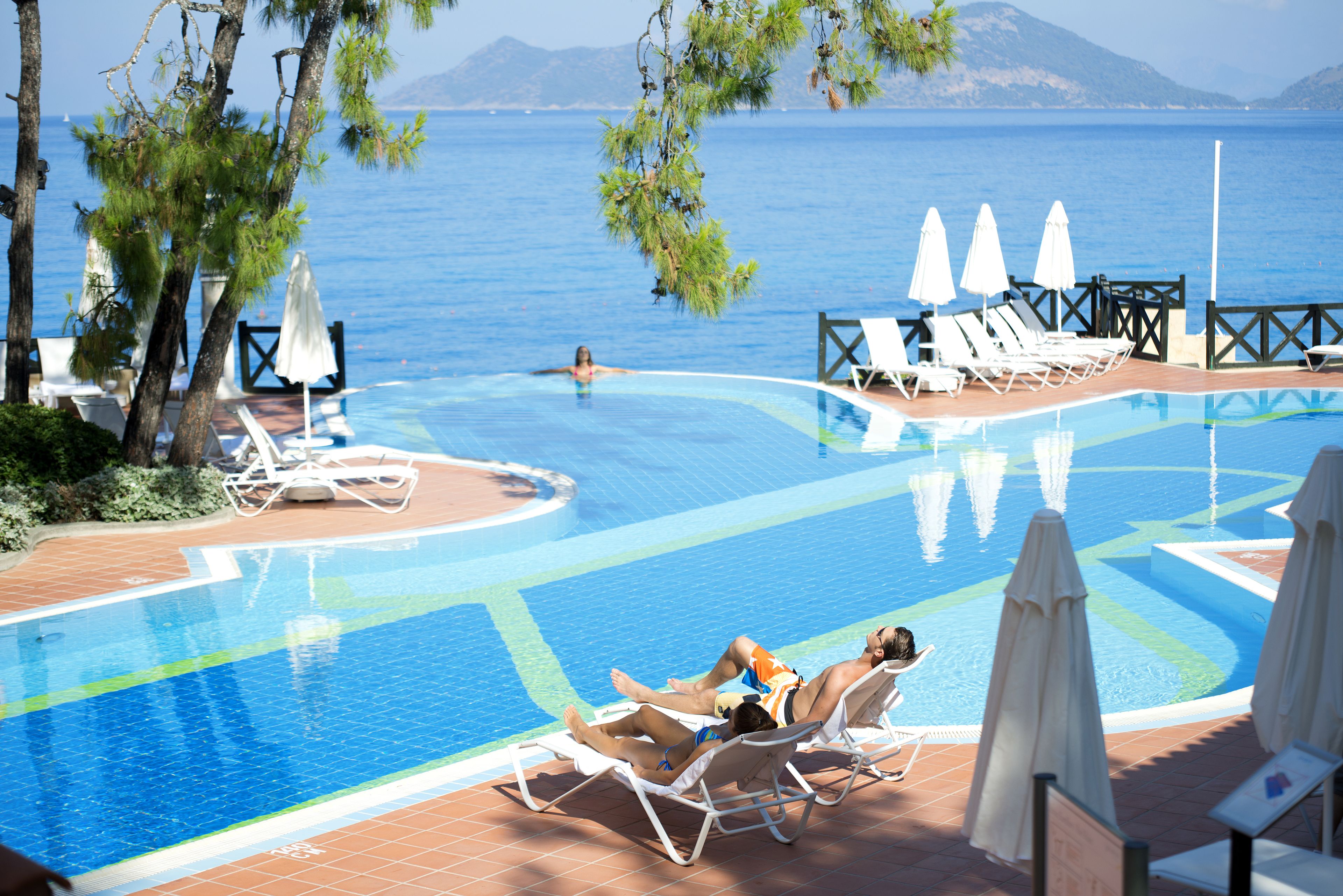 Piscina com espreguiçadeiras e guarda-sóis, rodeada por árvores e com vista para o mar.