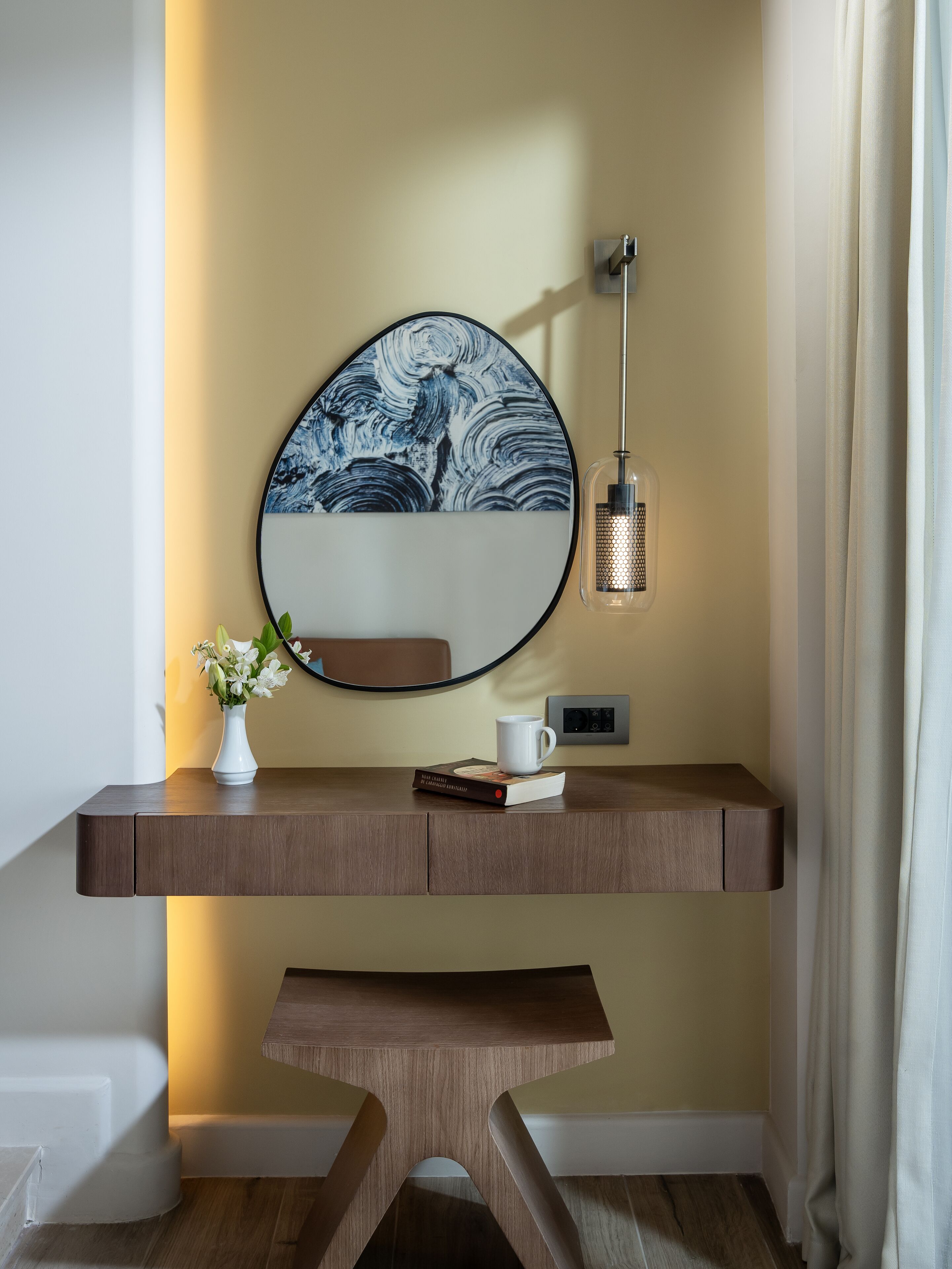 A modern desk with a round mirror, a vase with flowers, a book, and a cup.