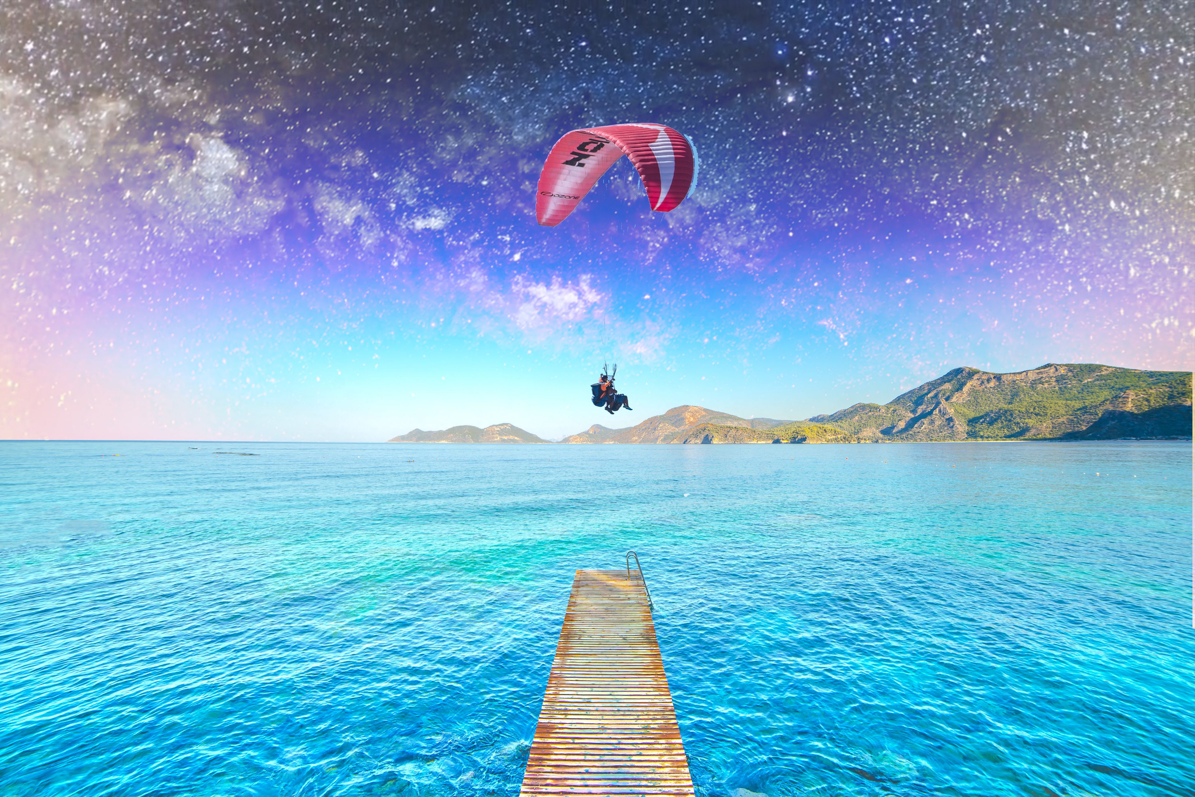 A clear view of a wooden pier extending into a calm sea, with a person paragliding in the distance.