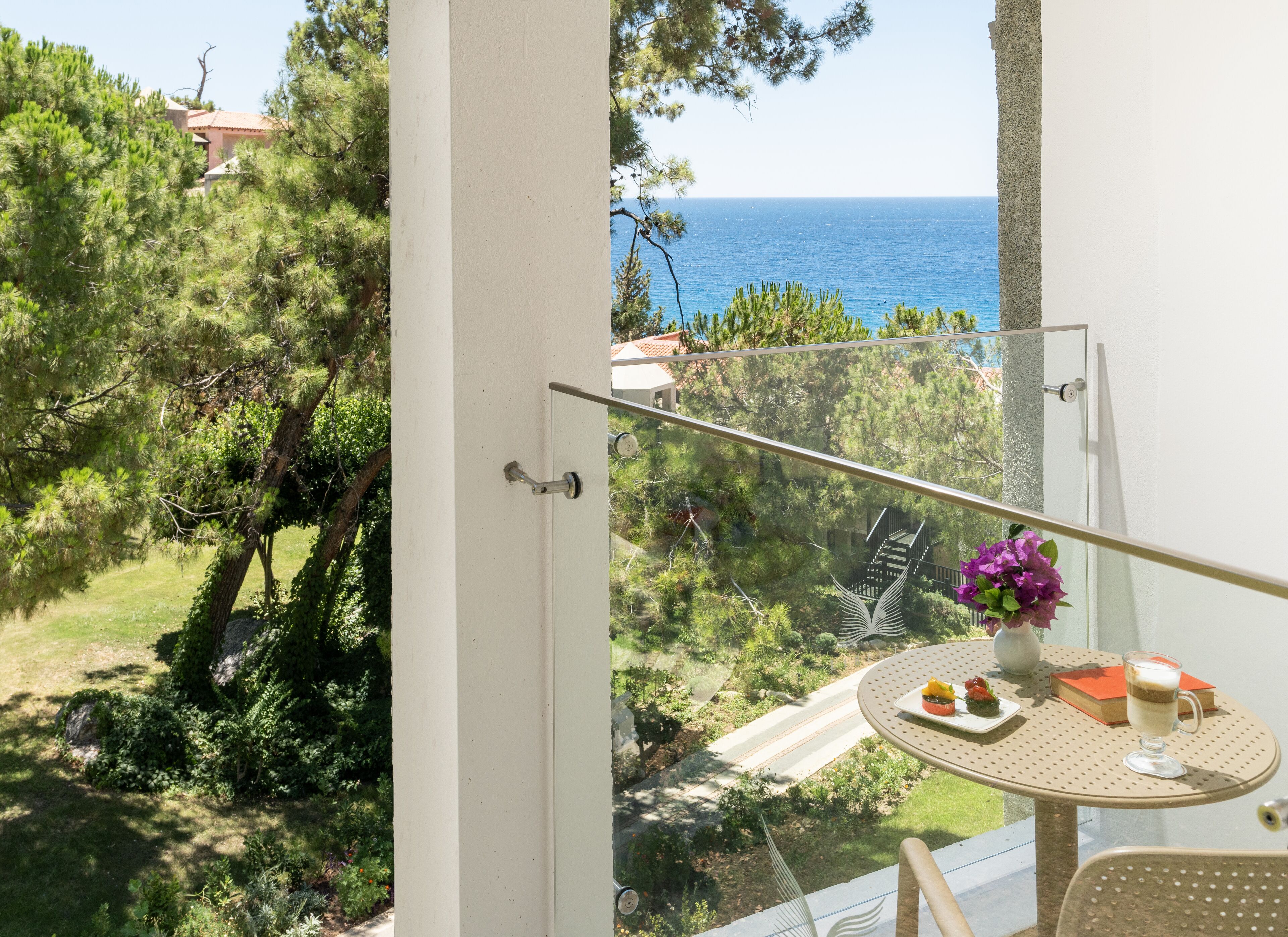 Ein Balkon mit Glasgeländer, der auf einen Garten und das Meer hinausblickt.