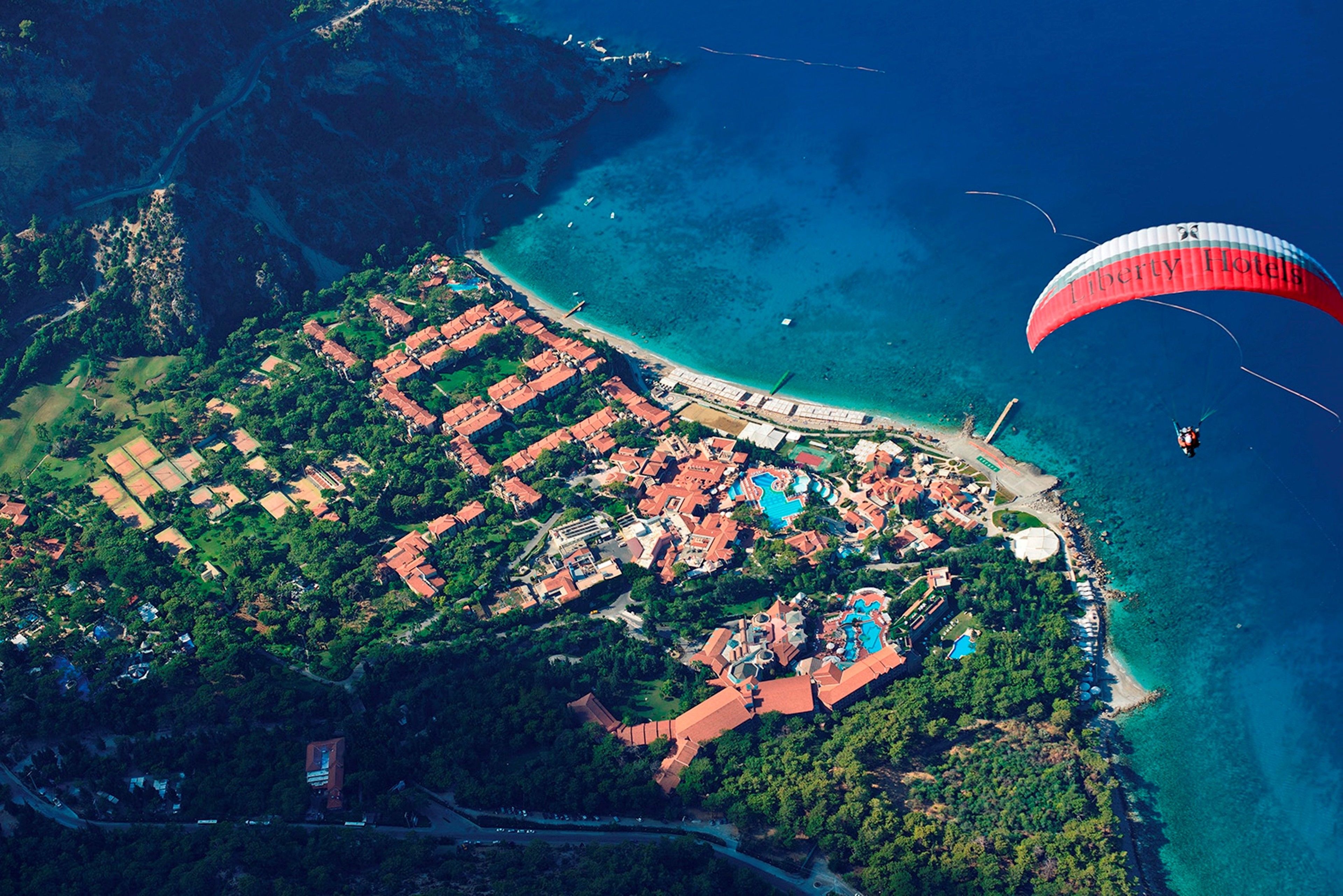 Vista aérea de un complejo turístico en la costa, con playa, piscinas y actividad de parasailing.