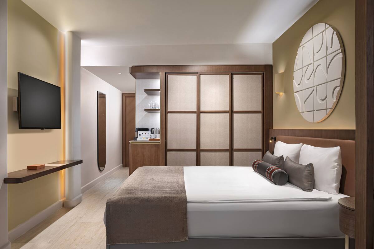 A modern hotel room with a large bed, a flat-screen TV, a wooden headboard, and a decorative wall piece.