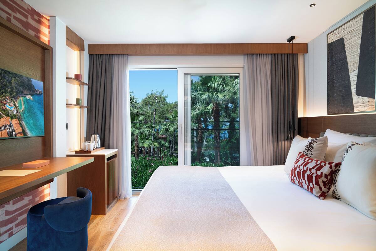 A modern hotel room with a large bed, a desk with a chair, a view of the outdoors, and a painting on the wall.