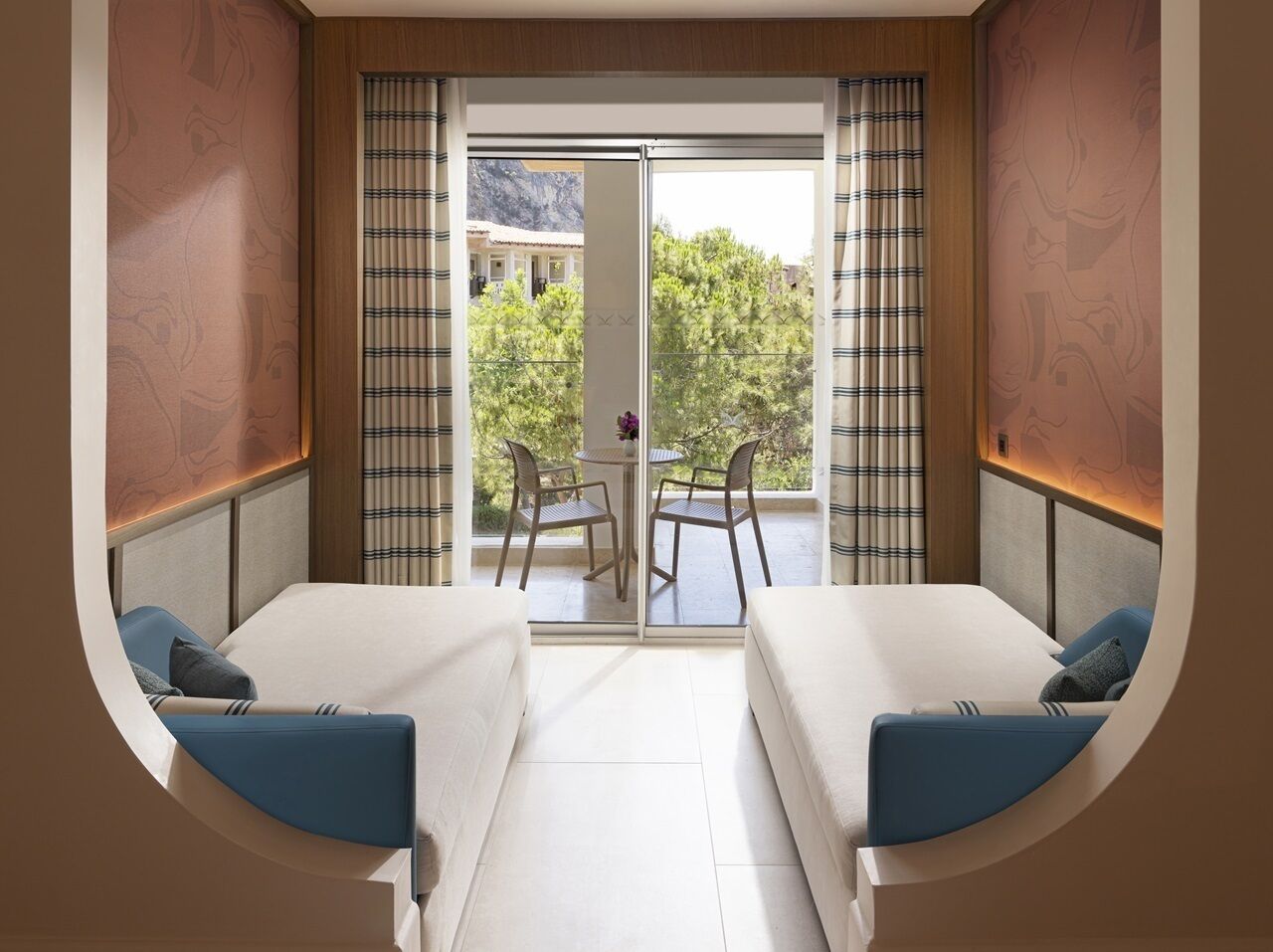 Ein Zimmer mit zwei Betten, einem kleinen Tisch mit Stühlen und Blick auf Grünflächen und Berge durch das Fenster.