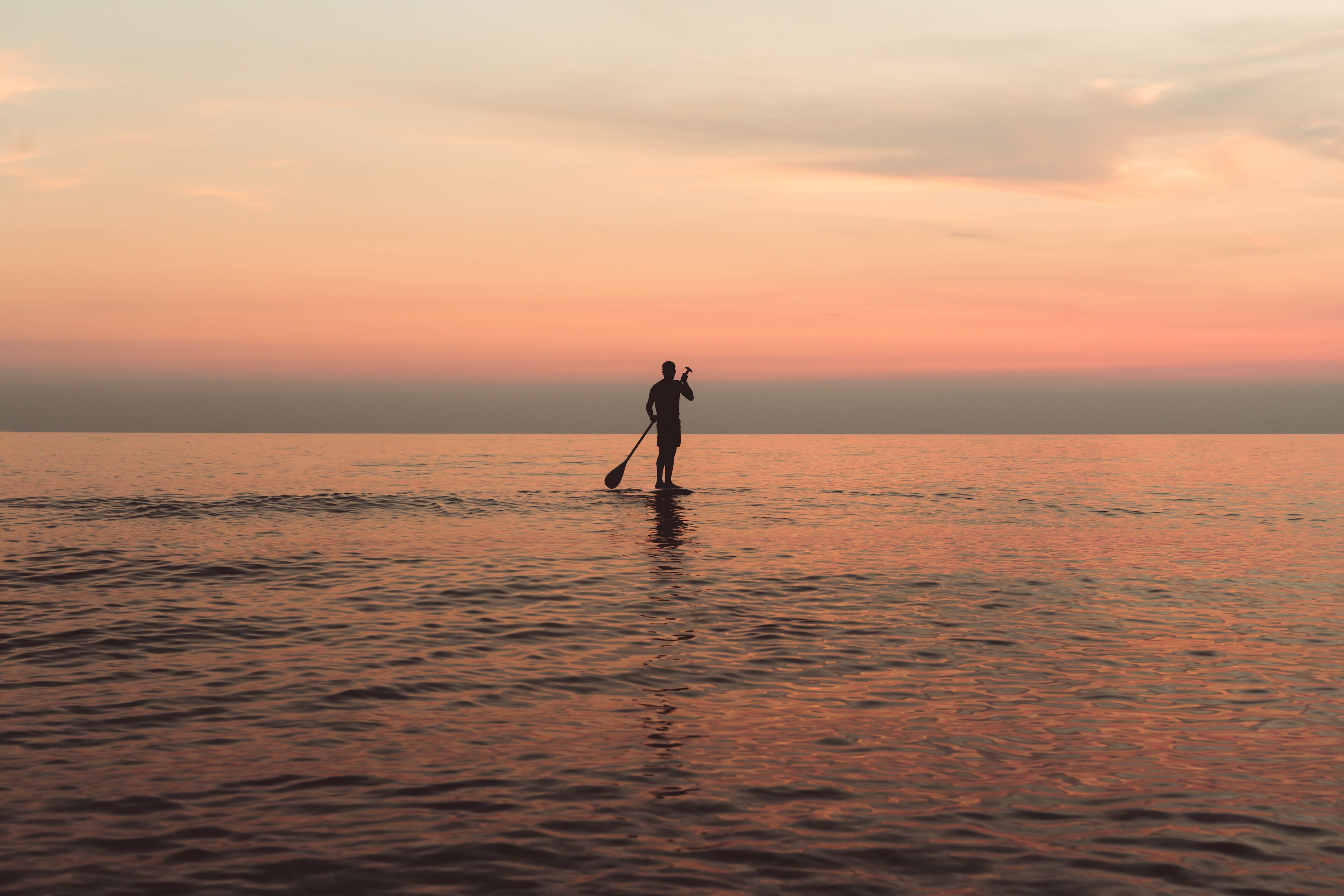 Una persona de pie en aguas poco profundas con una pala, probablemente en una playa durante el atardecer.
