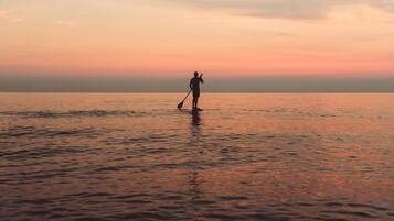 Una persona de pie en aguas poco profundas con una pala, probablemente en una playa durante el atardecer.