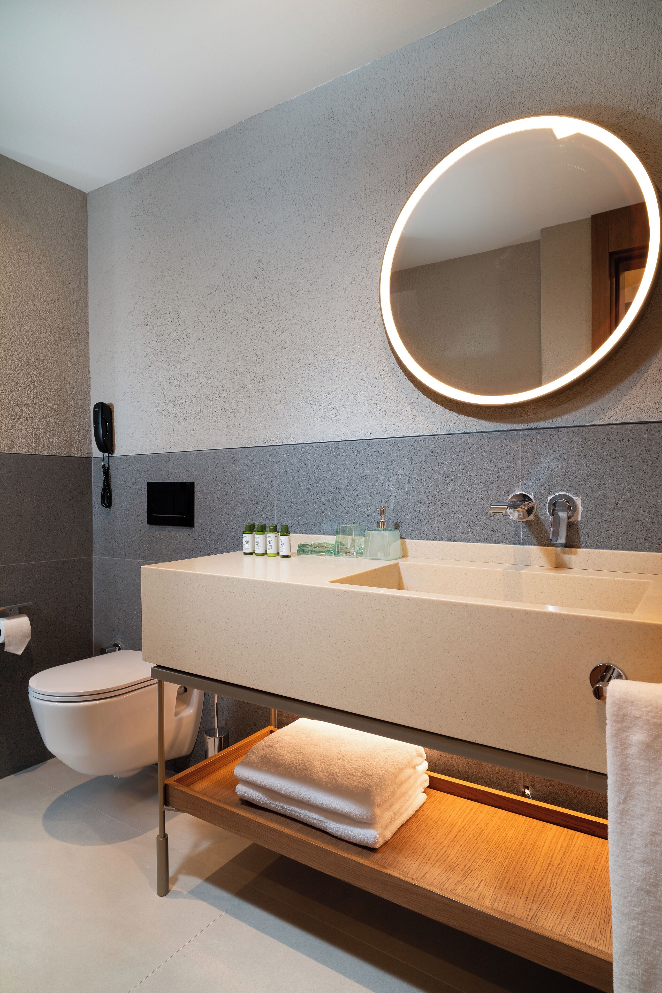 A modern bathroom with a large sink, a round mirror with a light, a toilet, and a shelf with towels.