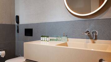 A modern bathroom with a large sink, a round mirror with a light, a toilet, and a shelf with towels.