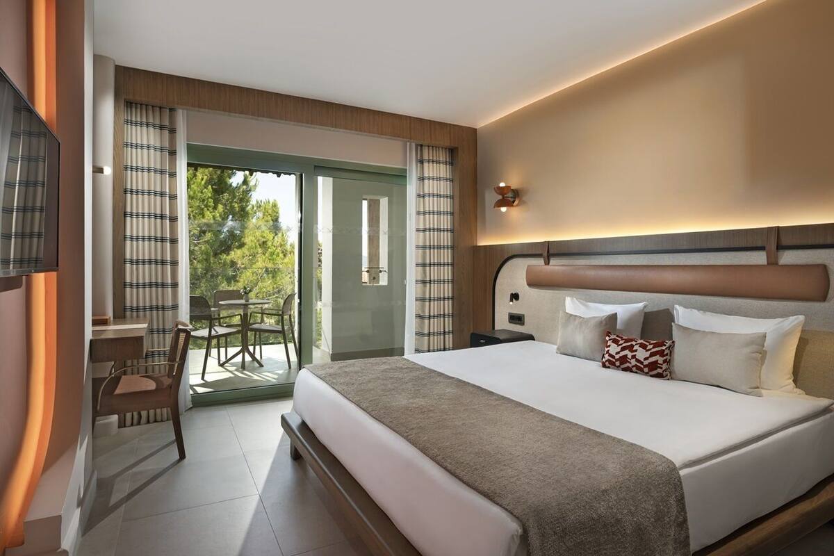 A modern hotel room with a large bed, a desk, and a chair. There is a balcony with a view of trees.