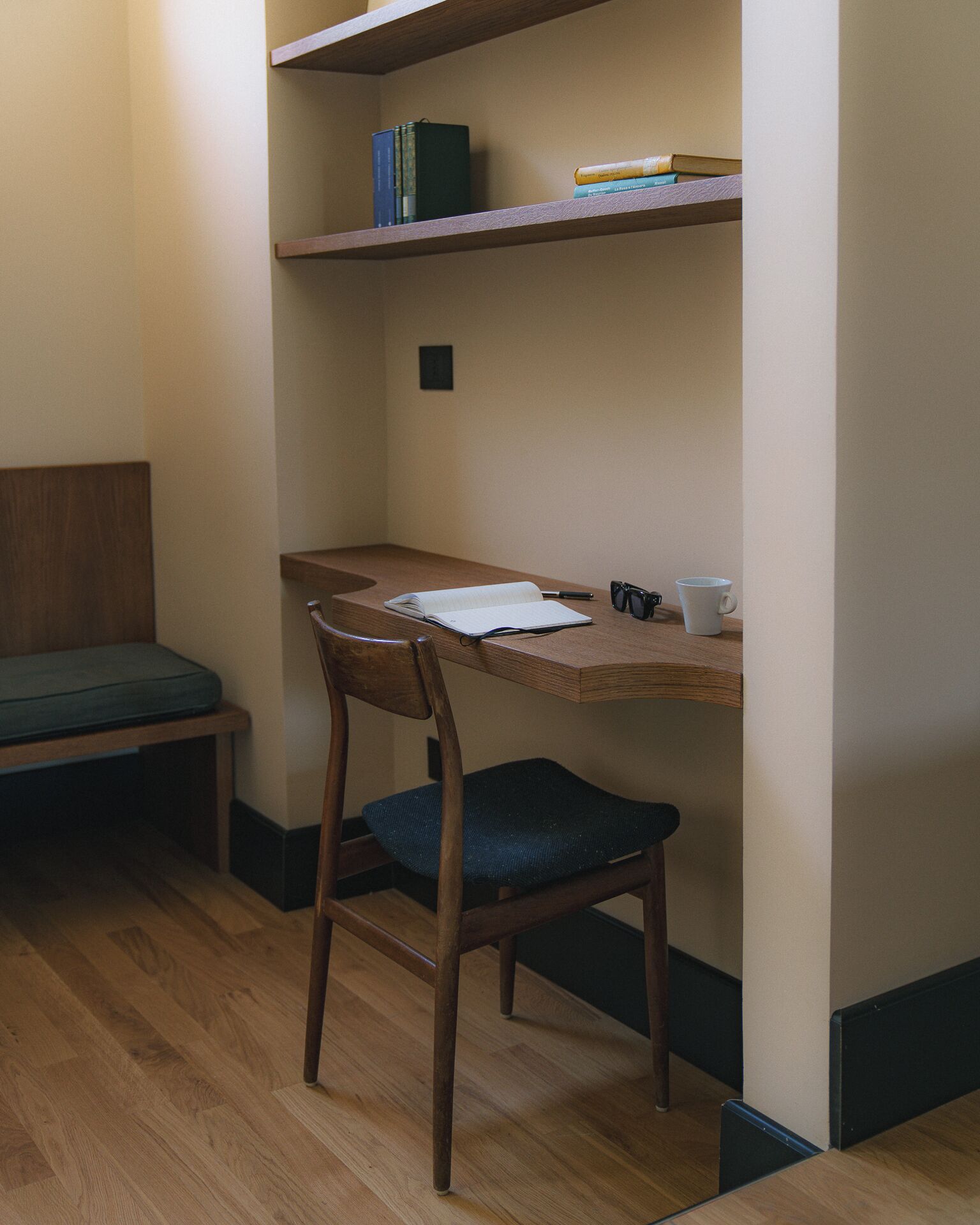 A small, cozy room with a wooden desk, a chair, a shelf with books, and a bench.