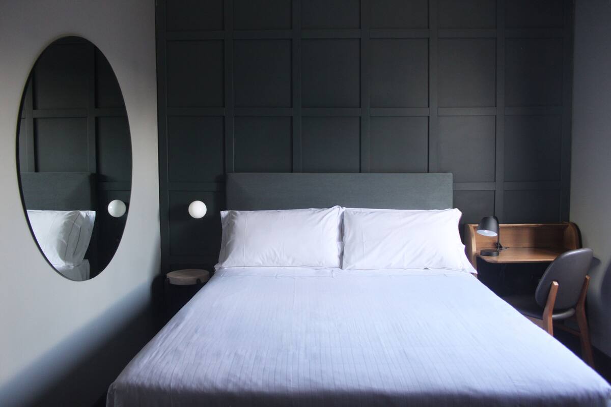 A neatly arranged hotel room with a large bed, a round mirror, a small desk with a chair, and a nightstand with a lamp.