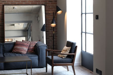A modern living room with a brick wall, a large mirror, a dark sofa, a wooden chair, and two black pendant lights.