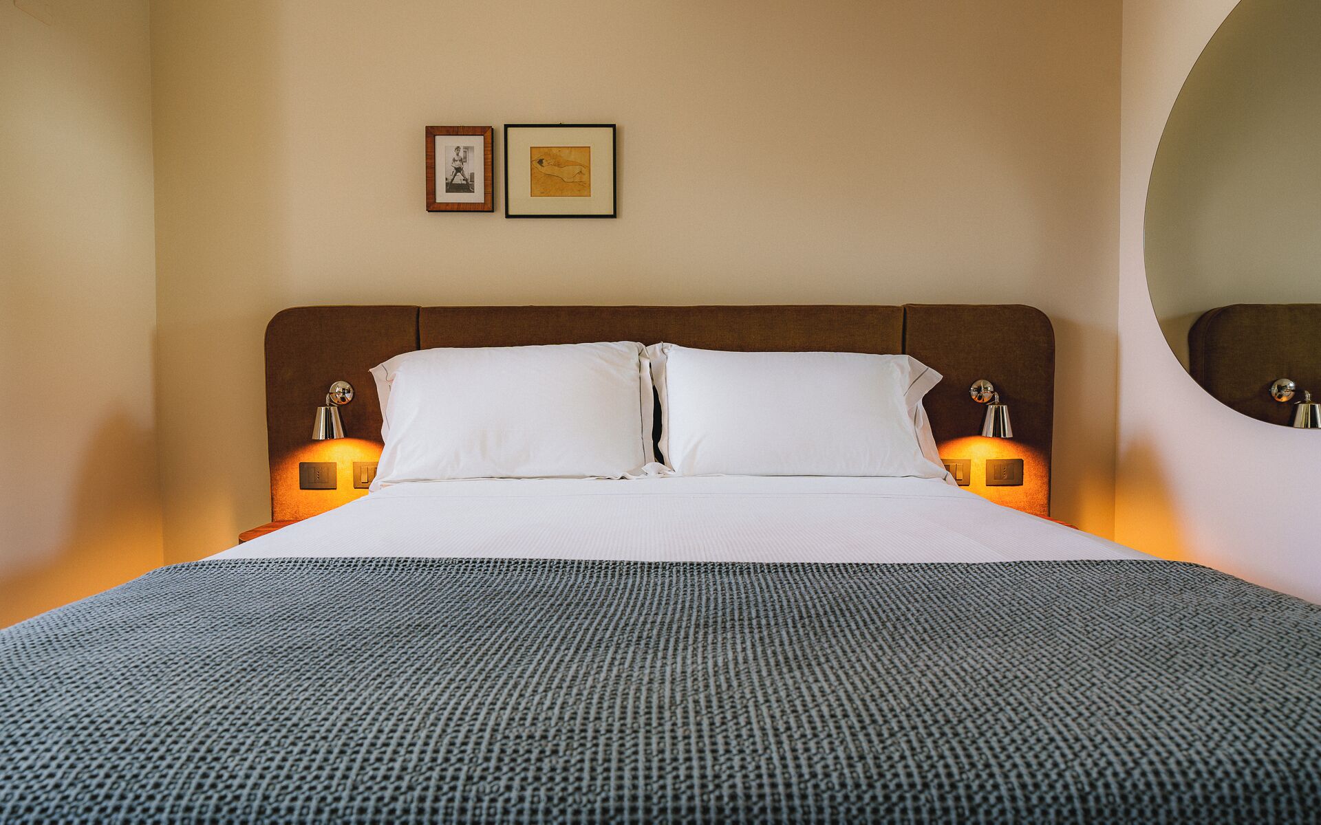 A neatly made bed with white linens and a gray textured blanket, flanked by two bedside lamps with round mirrors.