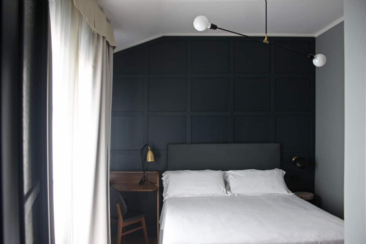 A bedroom with a bed, two bedside lamps, a desk with a chair, and a window with curtains.