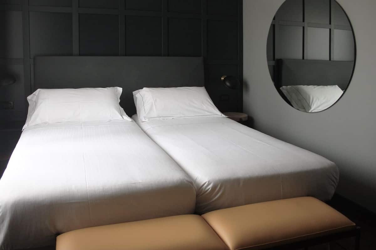 A double bed with white linens, a round mirror on the wall, and a bench at the foot of the bed.